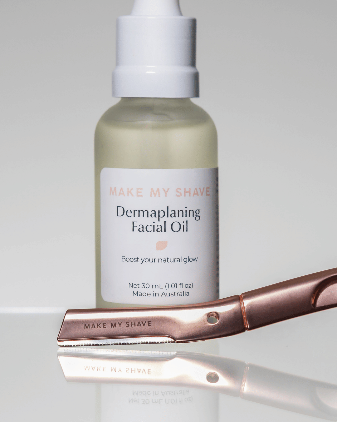 Bottle of 'Make My Shave' dermaplaning facial oil with a rose gold dermaplaning kit on a light grey background.