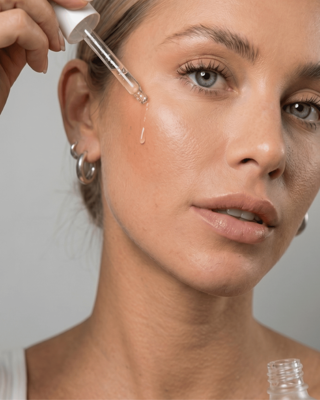 Close-up of a woman applying a dermaplaning facial oil via dropper to her cheek, with a single drop of liquid dripping onto her skin against a neutral grey background.