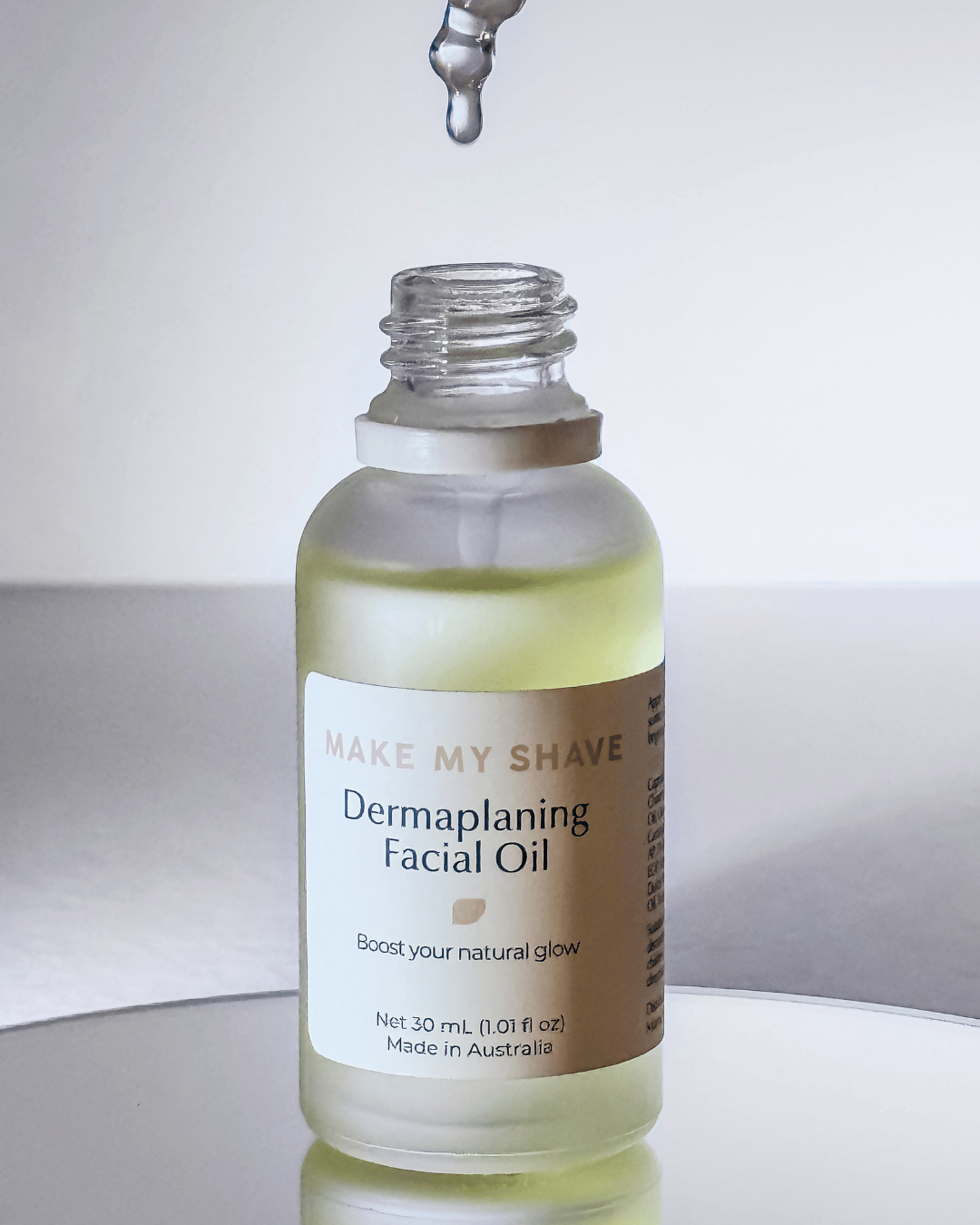 A 30 mL glass bottle of Make My Shave Dermaplaning Facial Oil with a white label, shown on a reflective surface with a dropper above it.