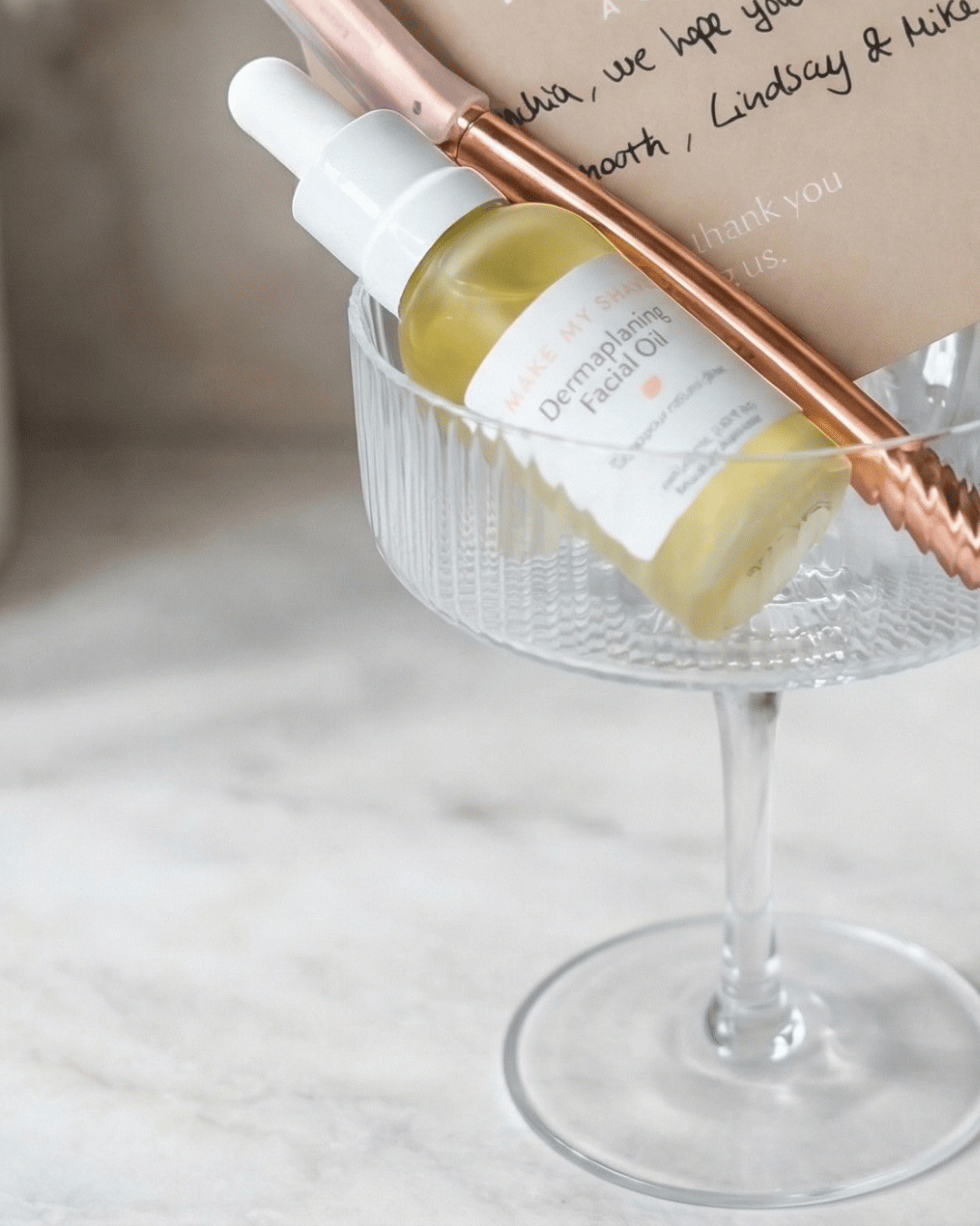 A small dropper bottle labeled 'Dermaplaning Facial Oil' in a clear glass bowl, styled with a rose gold metal spoon and a kraft paper note on a light marble surface. Oil is Australian made. 