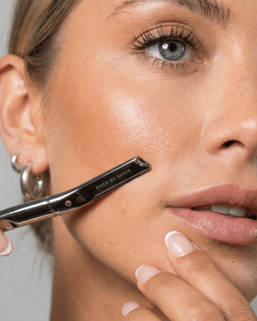 Close-up of a woman's face with the metal dermaplaning tool by Make My Shave against her cheek. 