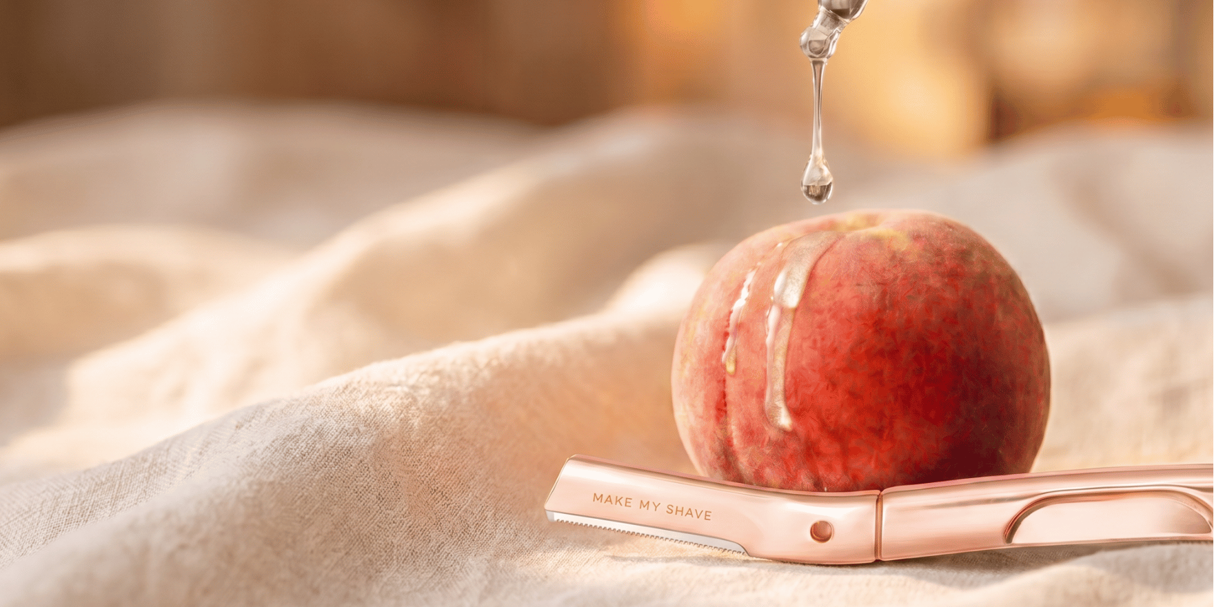 A rose gold dermaplaning facial razor lies on soft beige fabric, with a peach resting on top and a single water droplet hanging above it.