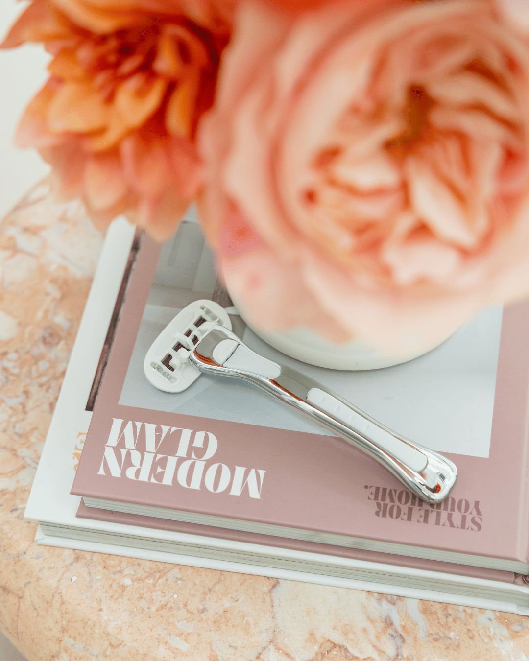 A reusable women's razor resting on a pink magazine, which is stacked on top of other books and partially covered by large peach-pink flowers in a white vase.